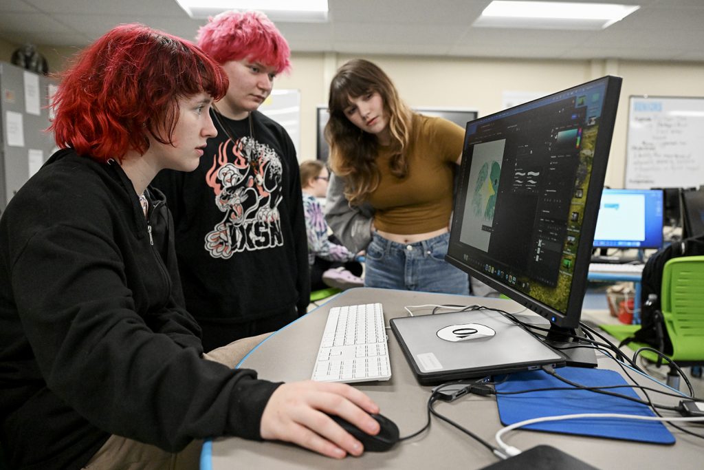 Three student peer onto a computer screen with digital art displayed.