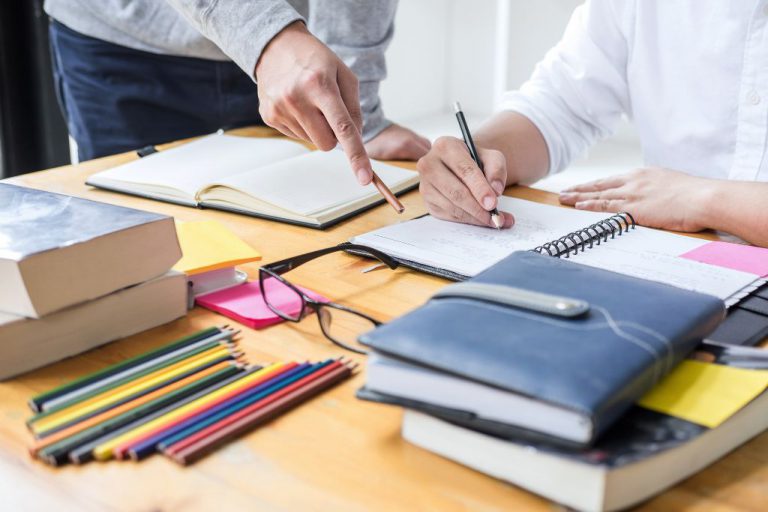 Closeup of hands of two people over a notebook with other books and pencils on the table.