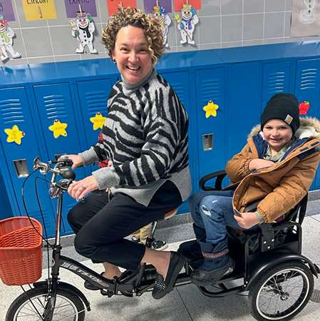 An elementary principal pedals a bike in the school hall with a student riding in a rear chair.
