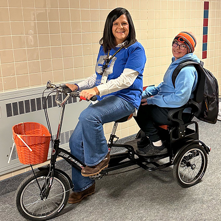 A principal pedals a bike in the school hall with a student riding in a rear chair.