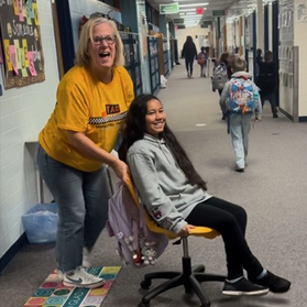 A principal wearing a t-shirt that says "principal taxi" mushes a student through the hallway in a rolling chair.