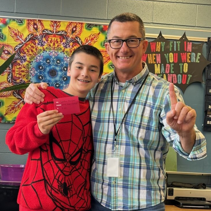 Scot Sargent stands next to a student, both happily raise one index finger in a classroom setting.